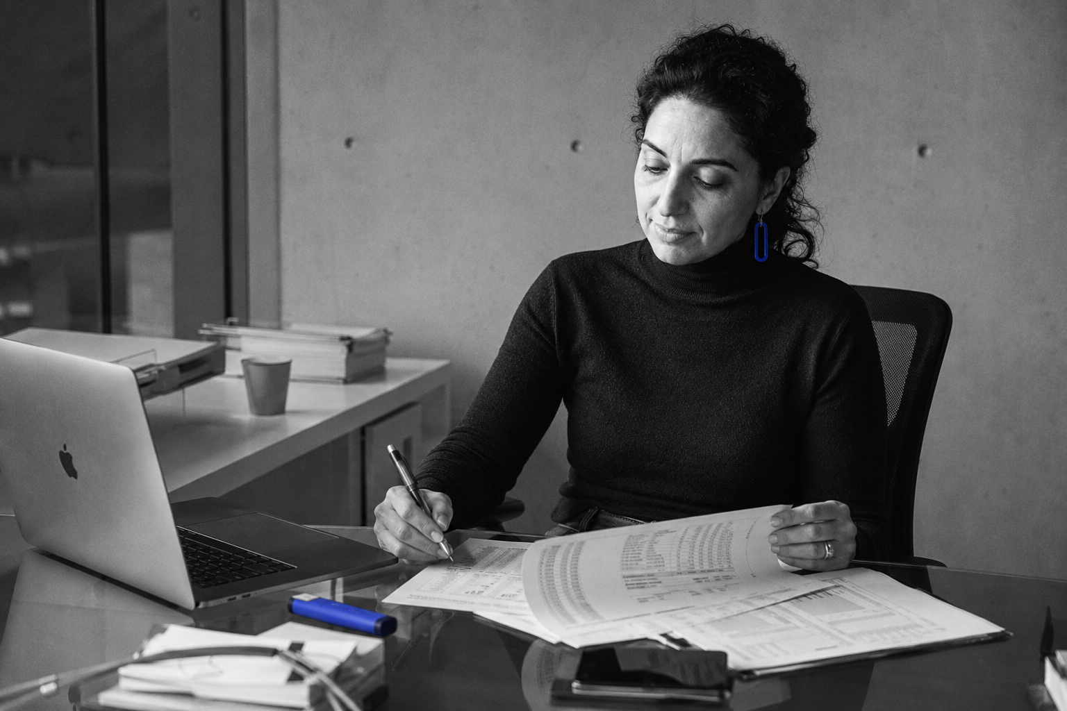 Petra Stavros at a clean glass desk reviewing P and L documents, single cobalt highlighter visible as the only colour pop, hard side light