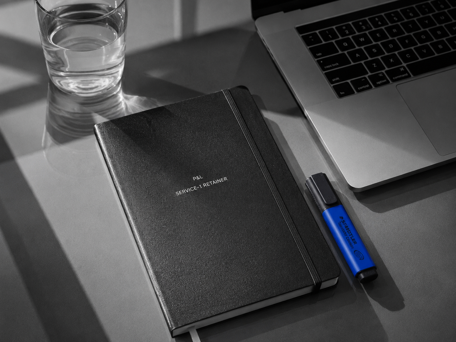 Petra's office: clean glass desk with hardcover notebook, MacBook Pro keyboard, single cobalt highlighter as only colour pop, hard side light