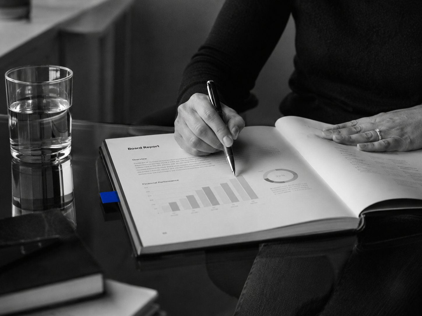 Glass conference table with single bound annual report, pen mid-annotation, cobalt-coloured tab marking a key page, hard side light