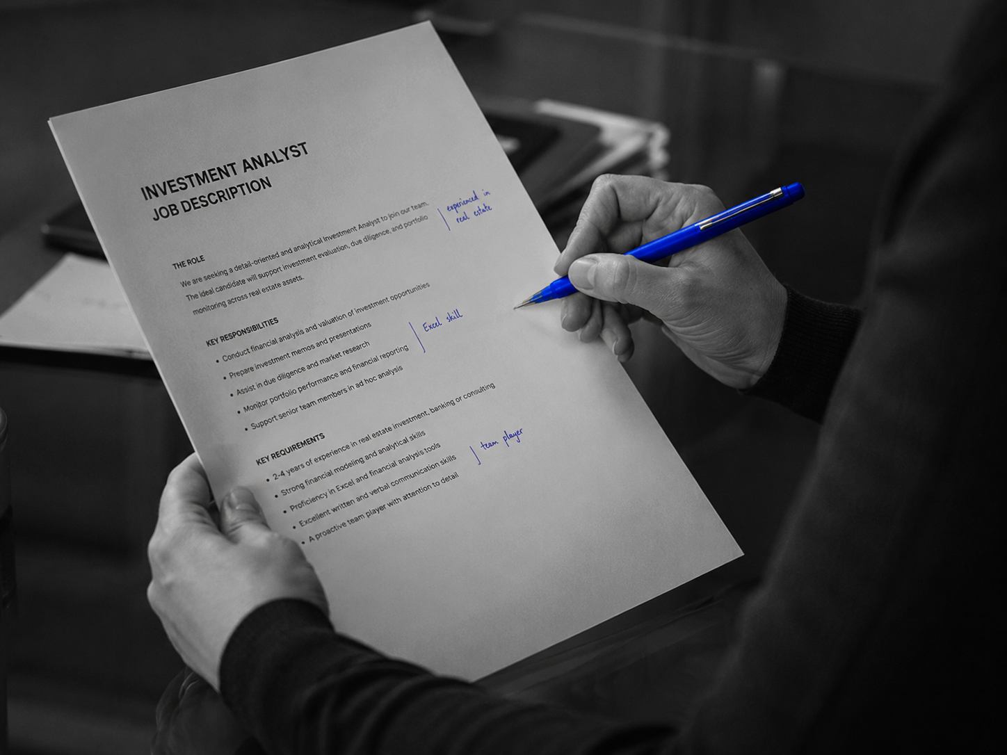 Hands holding a printed JD over glass desk, cobalt pen mid-margin annotation, hard side light