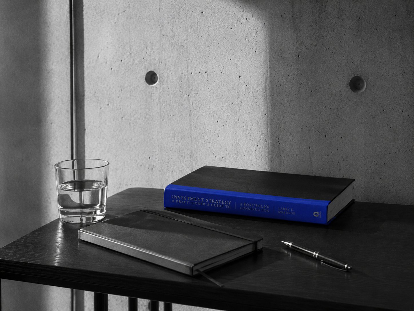Simple modern desk with a single book, notebook, pen and glass of water, concrete wall behind, cobalt book spine as only colour pop, hard side light